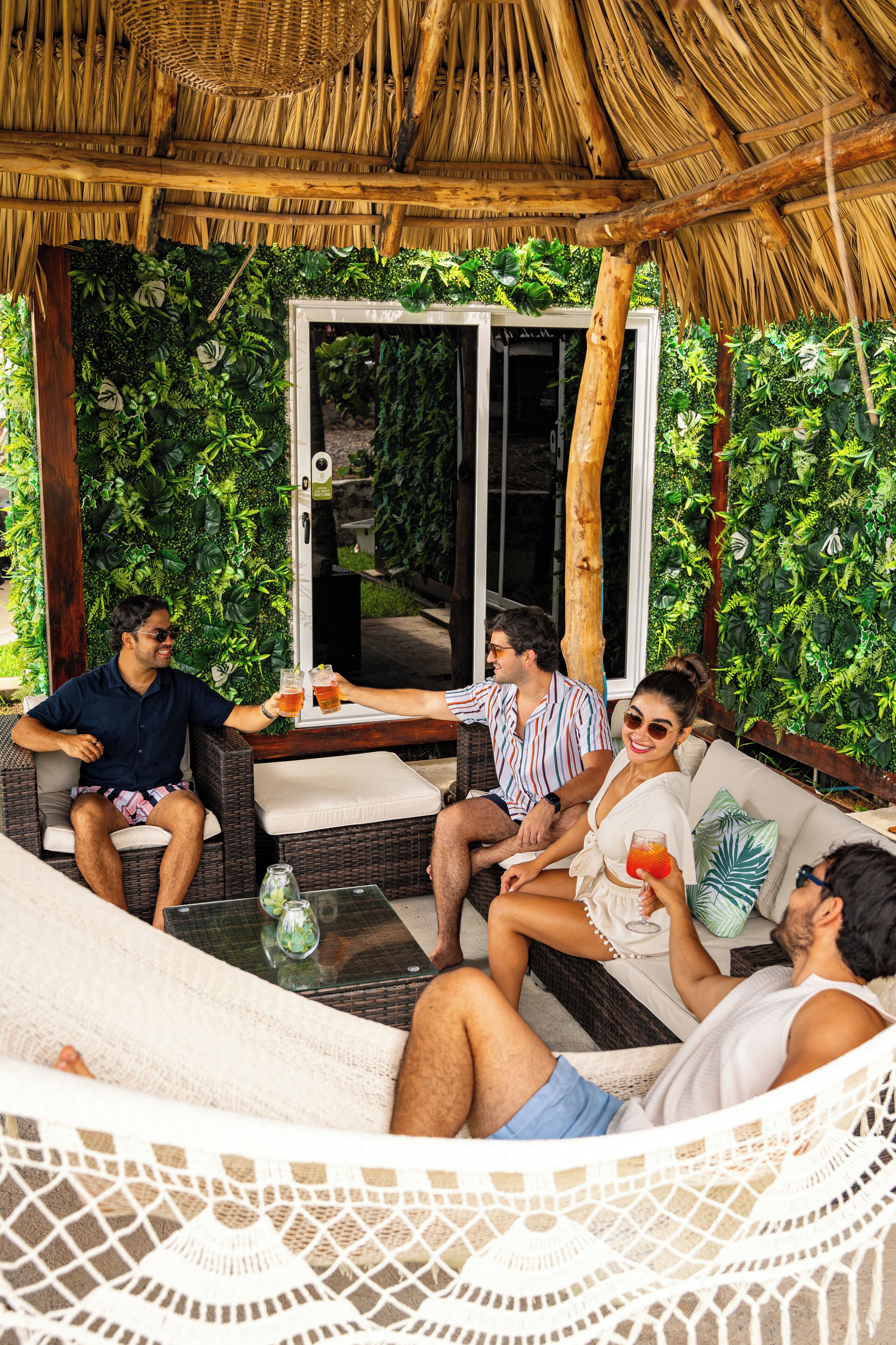 A group of people relaxing in a hammock and on a couch under a thatched roof, with lush greenery in the background.