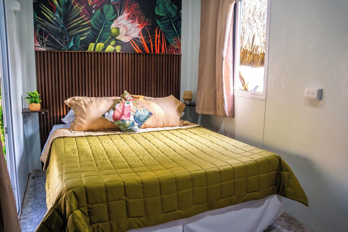 Una cama con colcha verde, dos almohadas, una mesita de noche con lámpara y una pared con un mural tropical.