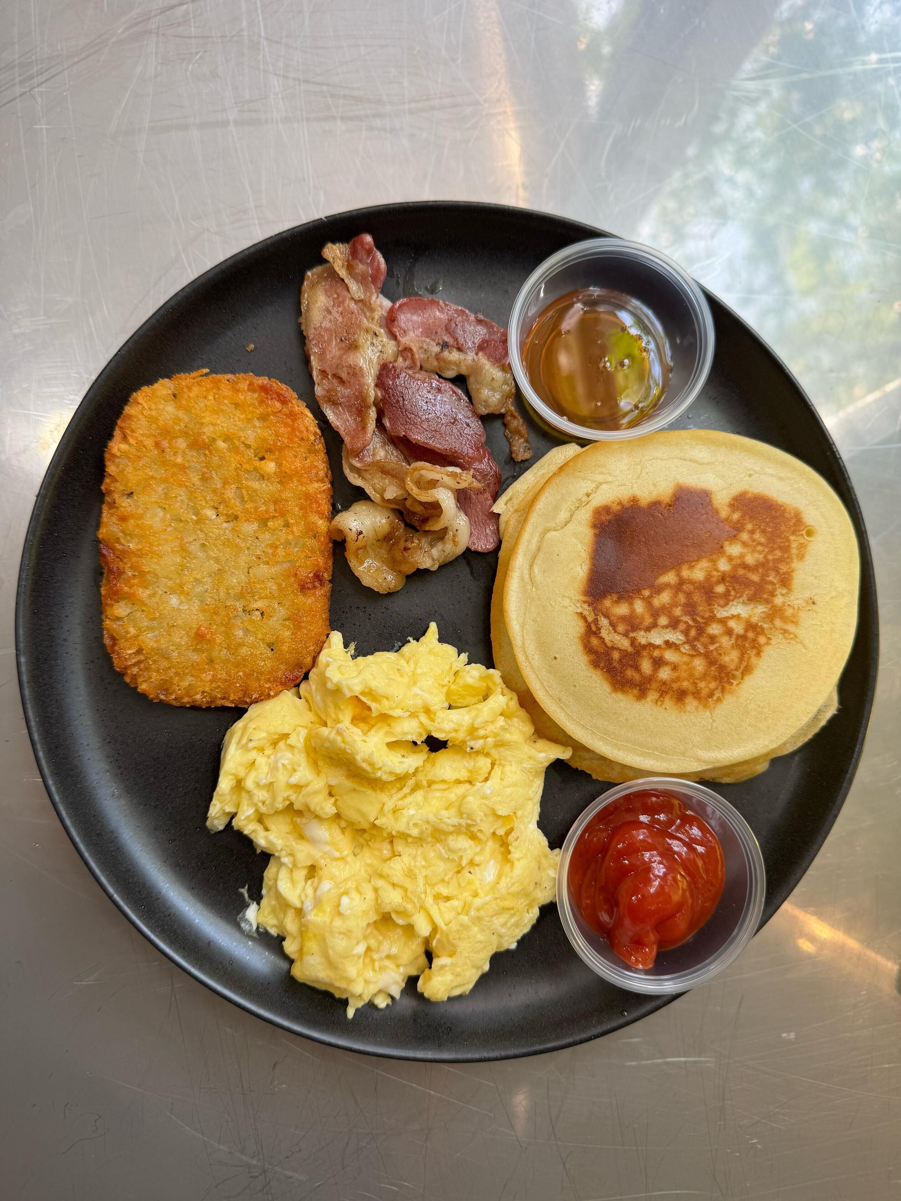 A breakfast plate with hash browns, bacon, scrambled eggs, pancakes, and ketchup.