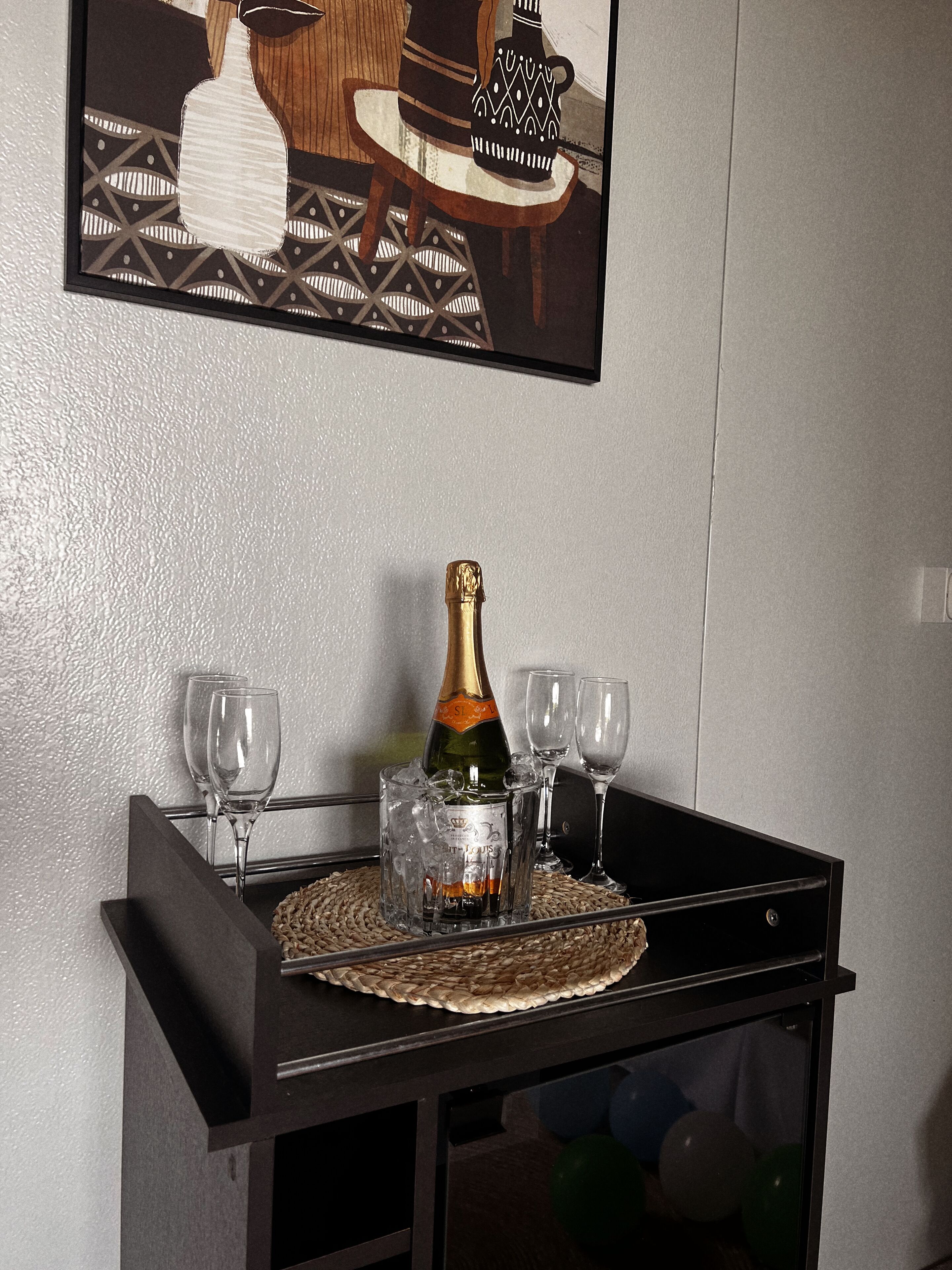 A bar area with a bottle of champagne, two glasses, and a woven placemat on a black table.
