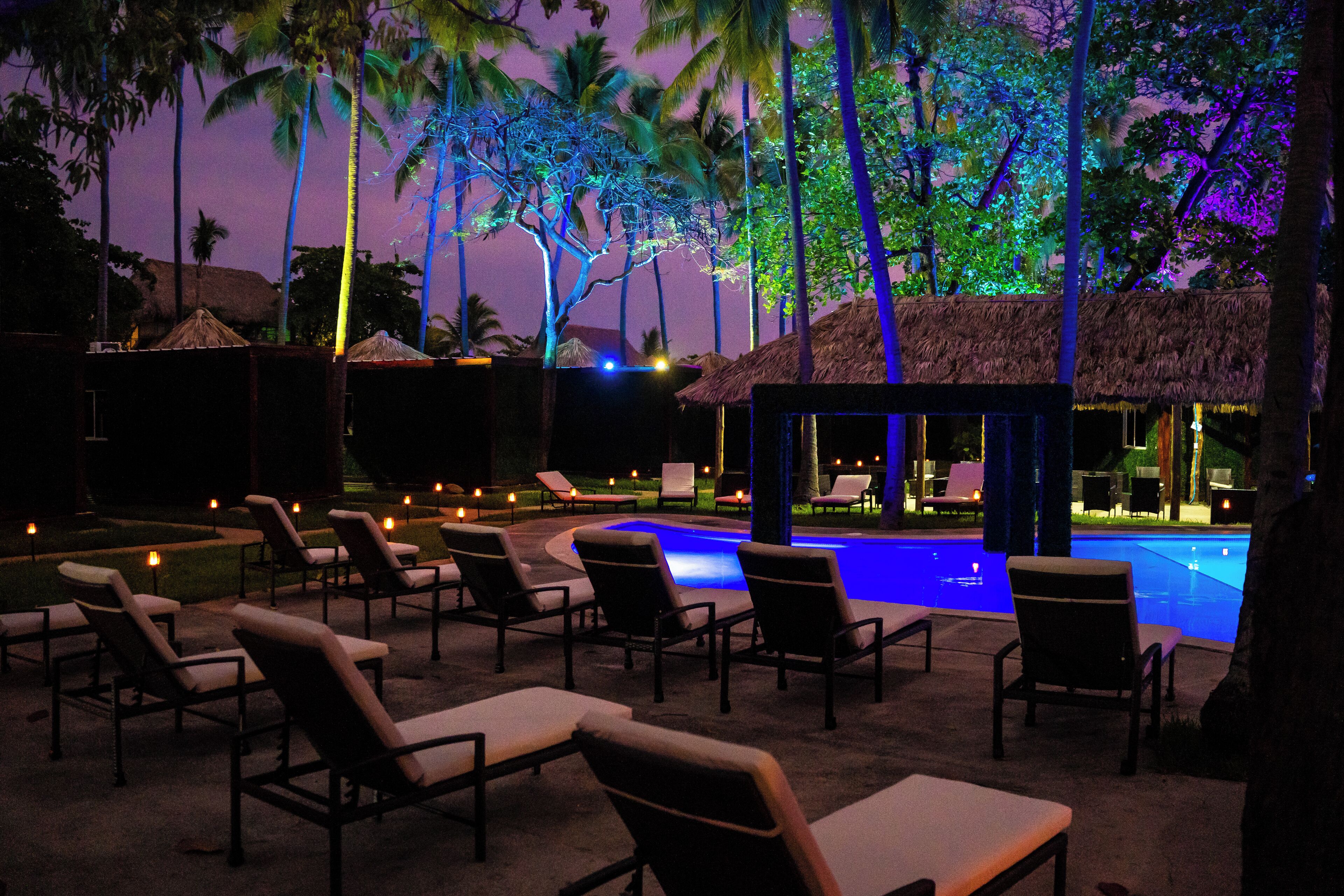 Un espace aménagé au bord de la piscine, avec des chaises longues et une structure à toit de chaume, le tout entouré de palmiers.