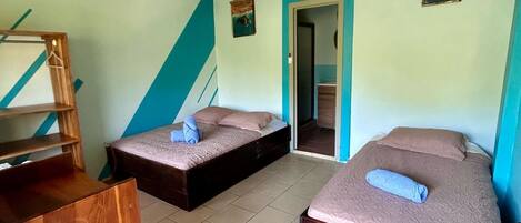 A small, compact room with two single beds, a wooden desk, and a door leading to another room.