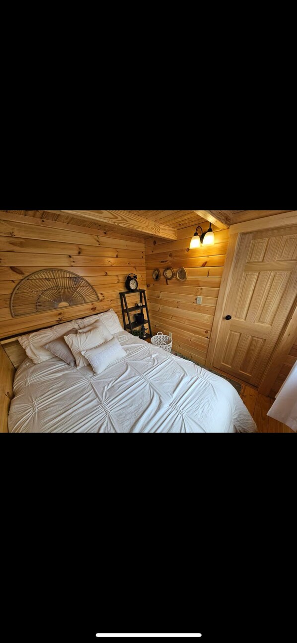 A wooden room with a bed, a wall fan, a clock, and a door.
