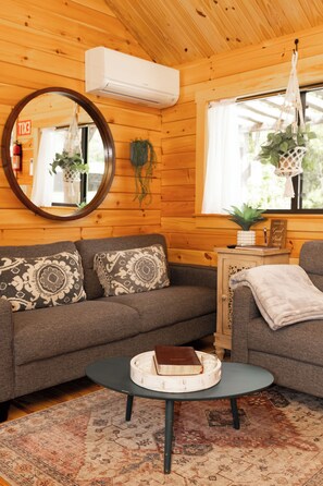 A cozy cabin interior with a wooden interior, a round mirror, a sofa with patterned cushions, a small table with a book and glass, and a hanging plant.