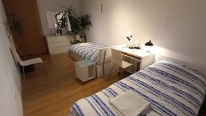Een slaapkamer met een bed, een bureau, een stoel en een klein tafeltje. Er hangt een spiegel aan de muur en op het bureau staat een plant.