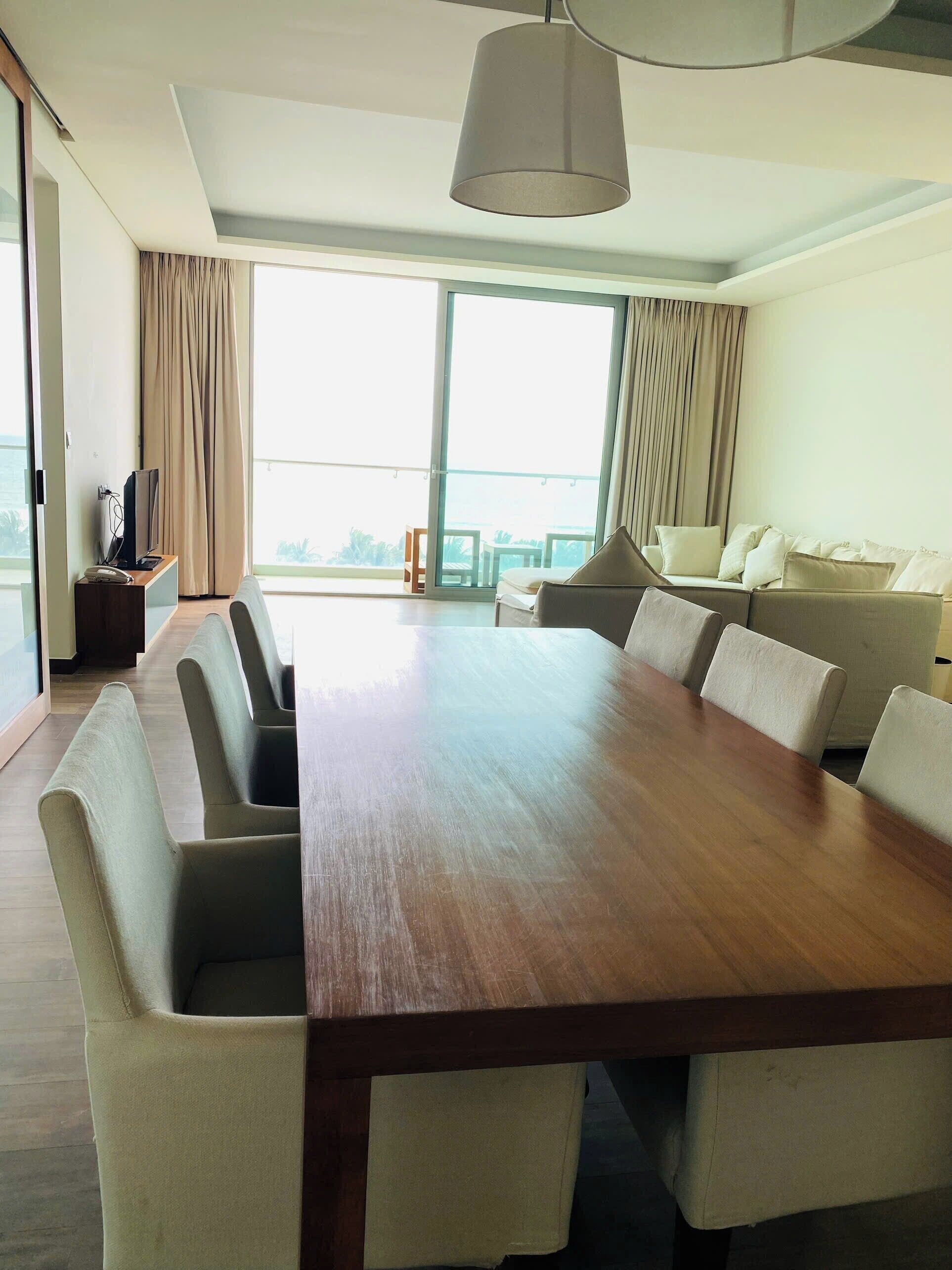 Uma sala de conferências com uma grande mesa de madeira, cadeiras estofadas bege, um sofá e uma televisão sobre um suporte.