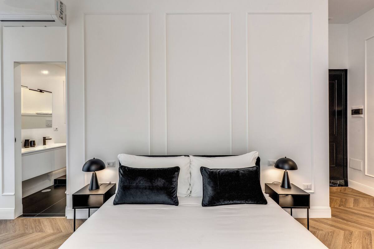 A modern bedroom with a large bed, two bedside tables with lamps, and a view into the bathroom.