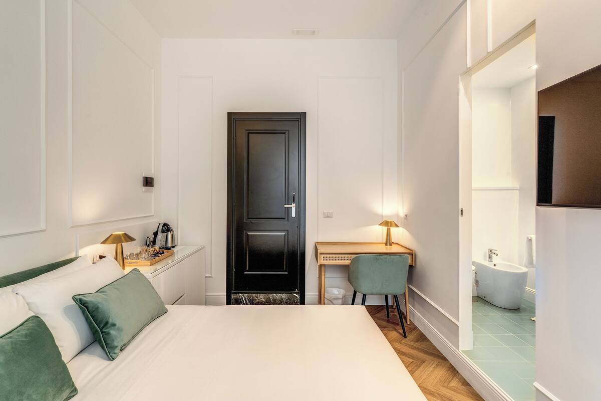 A hotel room with a bed, a desk, a chair, a black door, a bathroom with a bathtub, and a mirror.