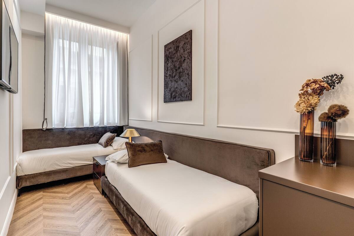 A modern hotel room with two beds, a large window with sheer curtains, a wall-mounted artwork, and a small table with decorative items.
