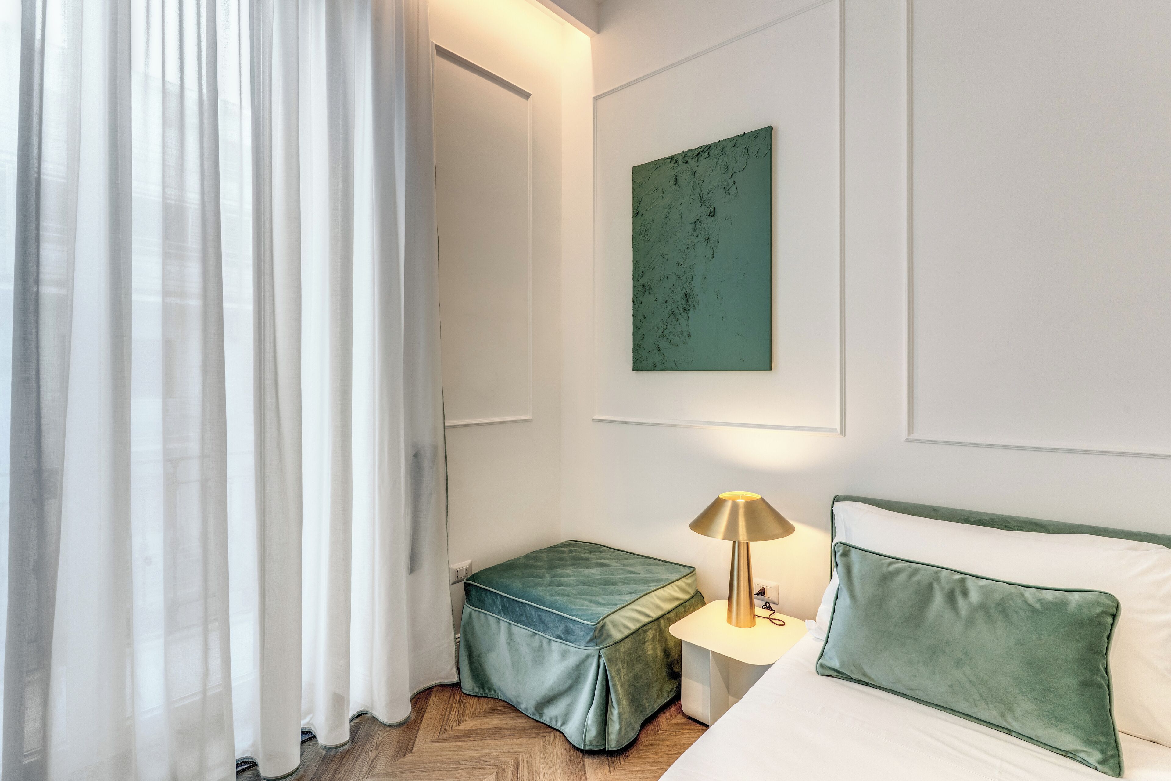 A hotel room with a bed, a nightstand with a lamp, a green ottoman, a green pillow, a green wall art piece, and sheer curtains.
