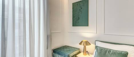 A hotel room with a bed, a nightstand with a lamp, a green ottoman, a green pillow, a green wall art piece, and sheer curtains.