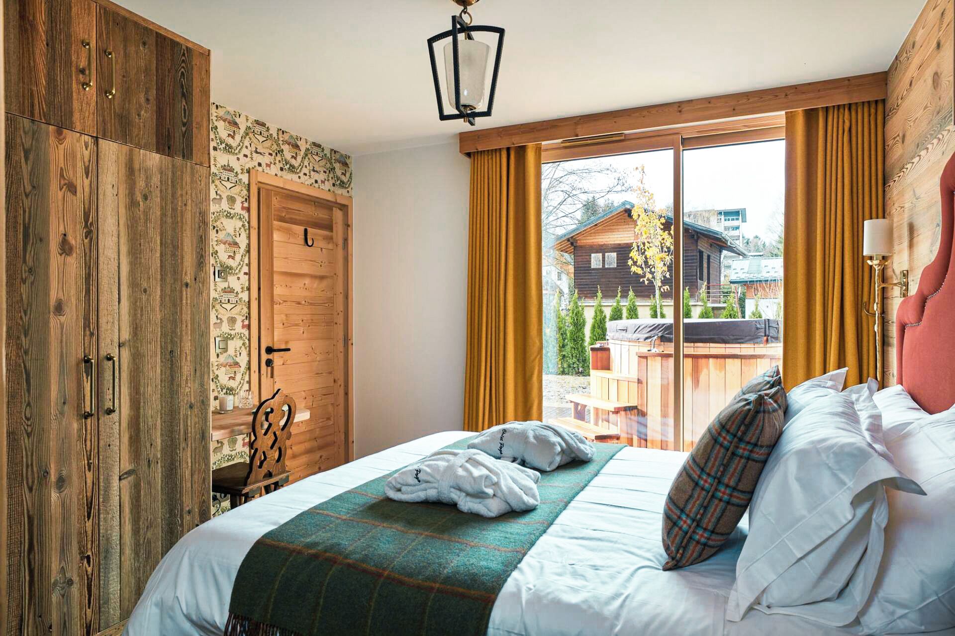 Une chambre avec une armoire en bois, un lit recouvert de draps blancs, une couverture à carreaux verts et une vue sur un chalet en bois par une grande fenêtre.