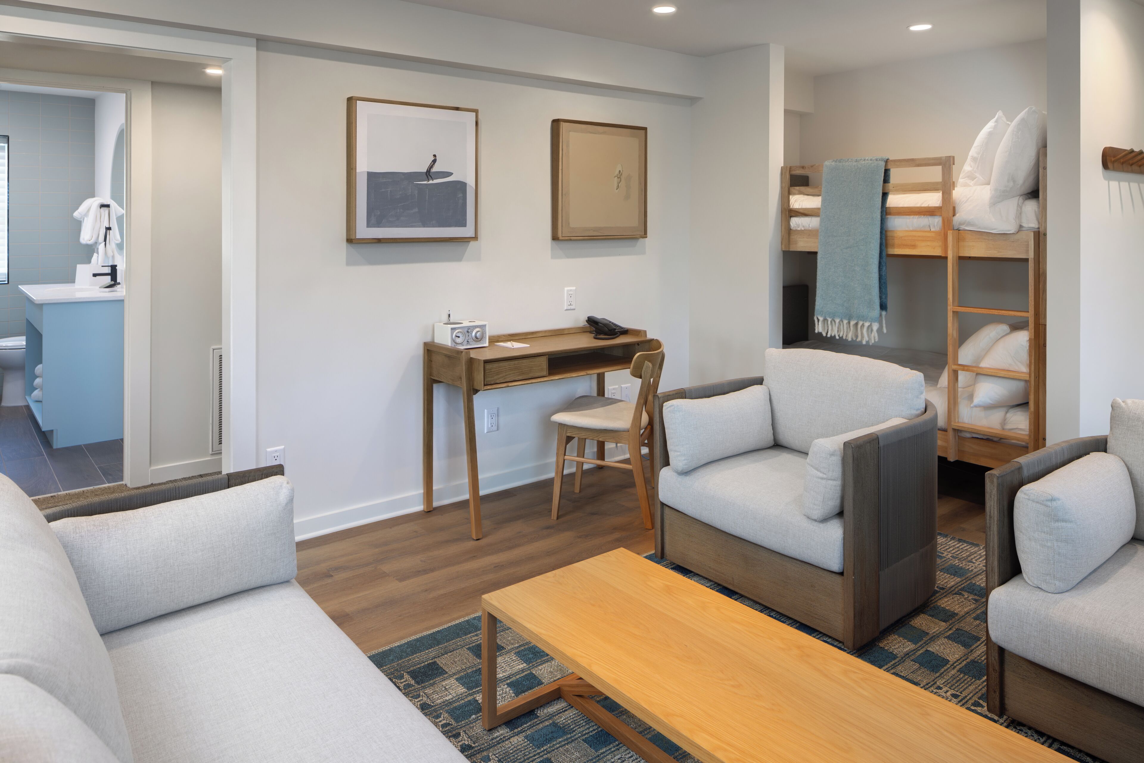 A modern, well-lit room with a sofa, a small wooden table, a desk with a chair, and a bunk bed with pillows.