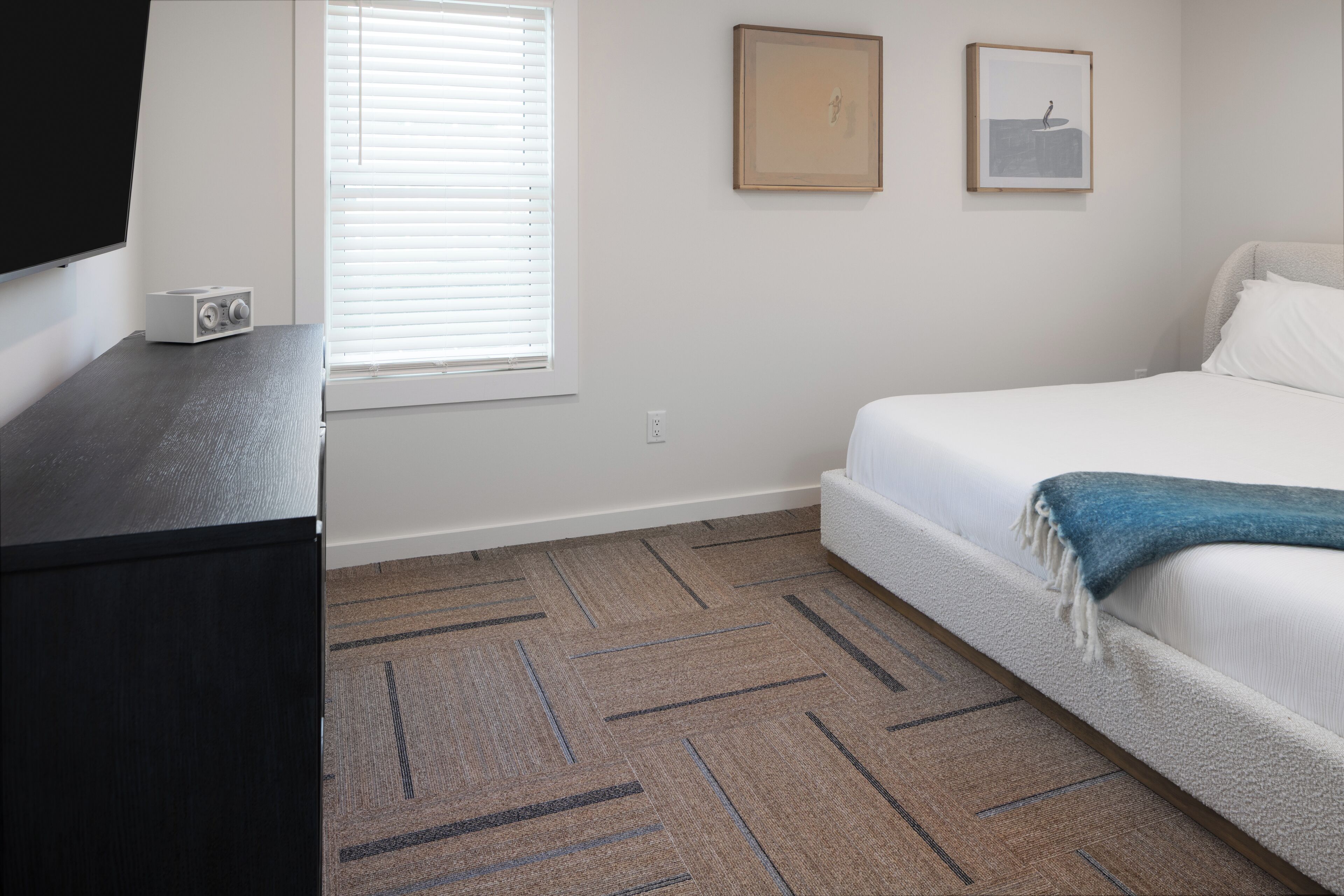A bedroom with a bed, a TV on a stand, a window with blinds, and a carpeted floor.