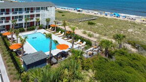 A pool area with lounge chairs and umbrellas, a beach with umbrellas and people, and a building with balconies. - Oceaneer (Carolina Beach)