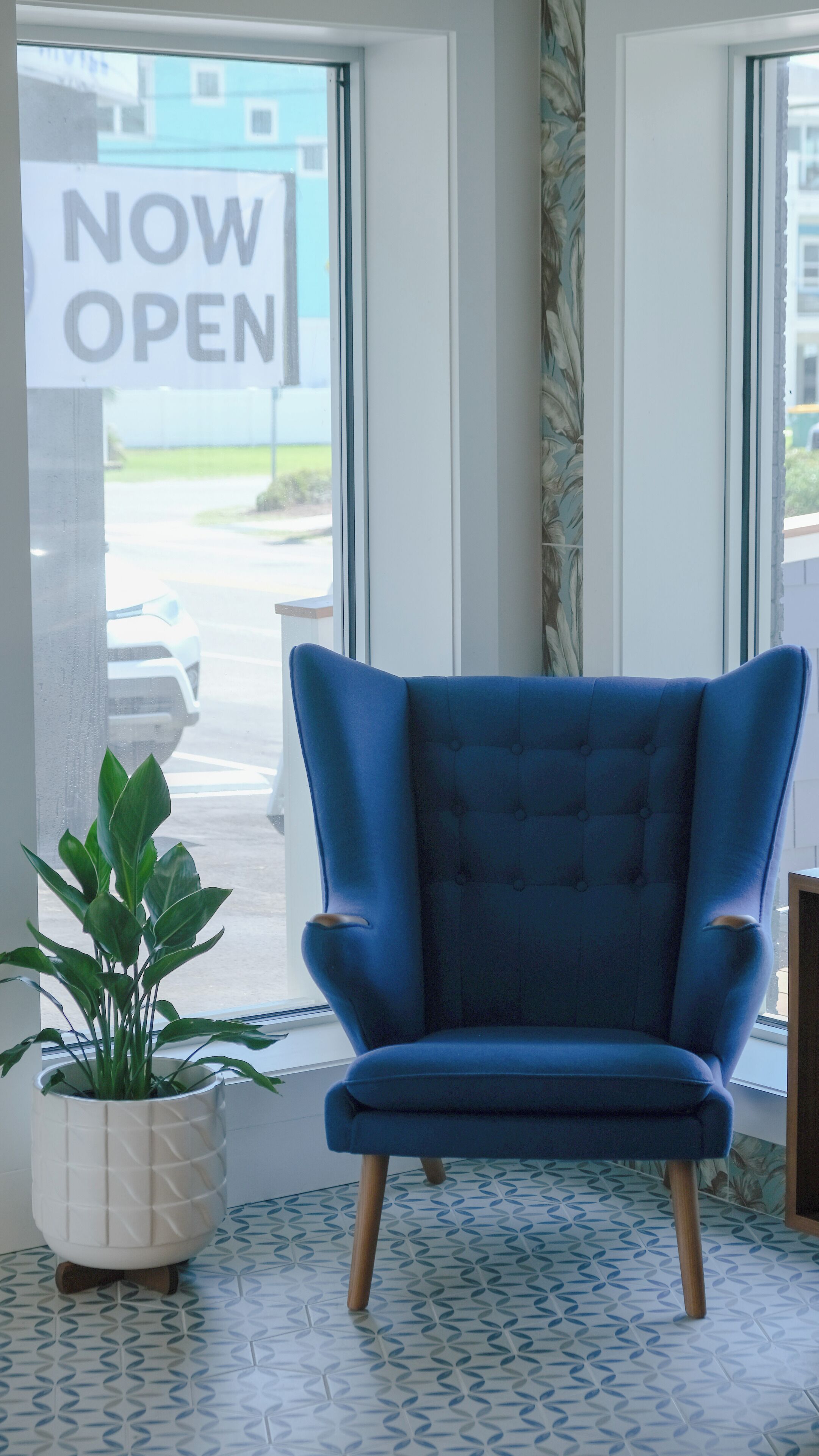 Una poltrona blu in una stanza con un'ampia finestra, una pianta in vaso e un cartello con la scritta 'NOW OPEN' visibile attraverso la finestra.