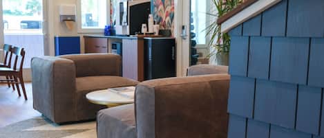 A modern living room with two brown sofas, a small round table, and a kitchen area with a trash bin and a refrigerator.