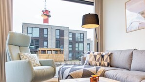 Ein modernes Wohnzimmer mit einer Couch, einem Sessel und einem Couchtisch. Ein Fenster bietet Blick auf ein Gebäude und einen Turm.