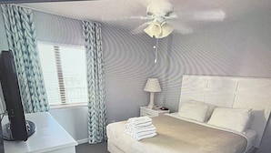Habitación de hotel con cama, escritorio, ordenador, lámpara y ventana con cortinas.
