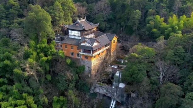 Un bâtiment traditionnel doté d’un toit à plusieurs niveaux, entouré d’une forêt dense et d’une petite cascade.