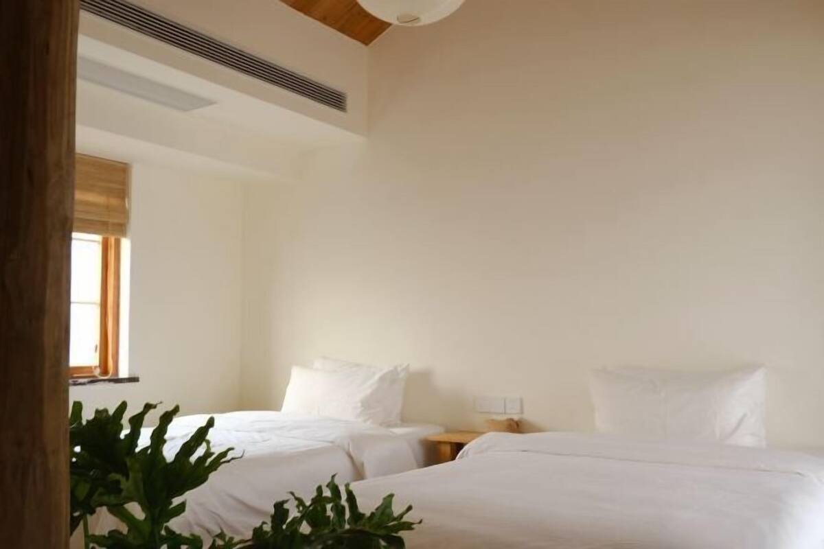 Een hotelkamer met twee bedden, een raam met zonwering, een plant en een ventilatie rooster.