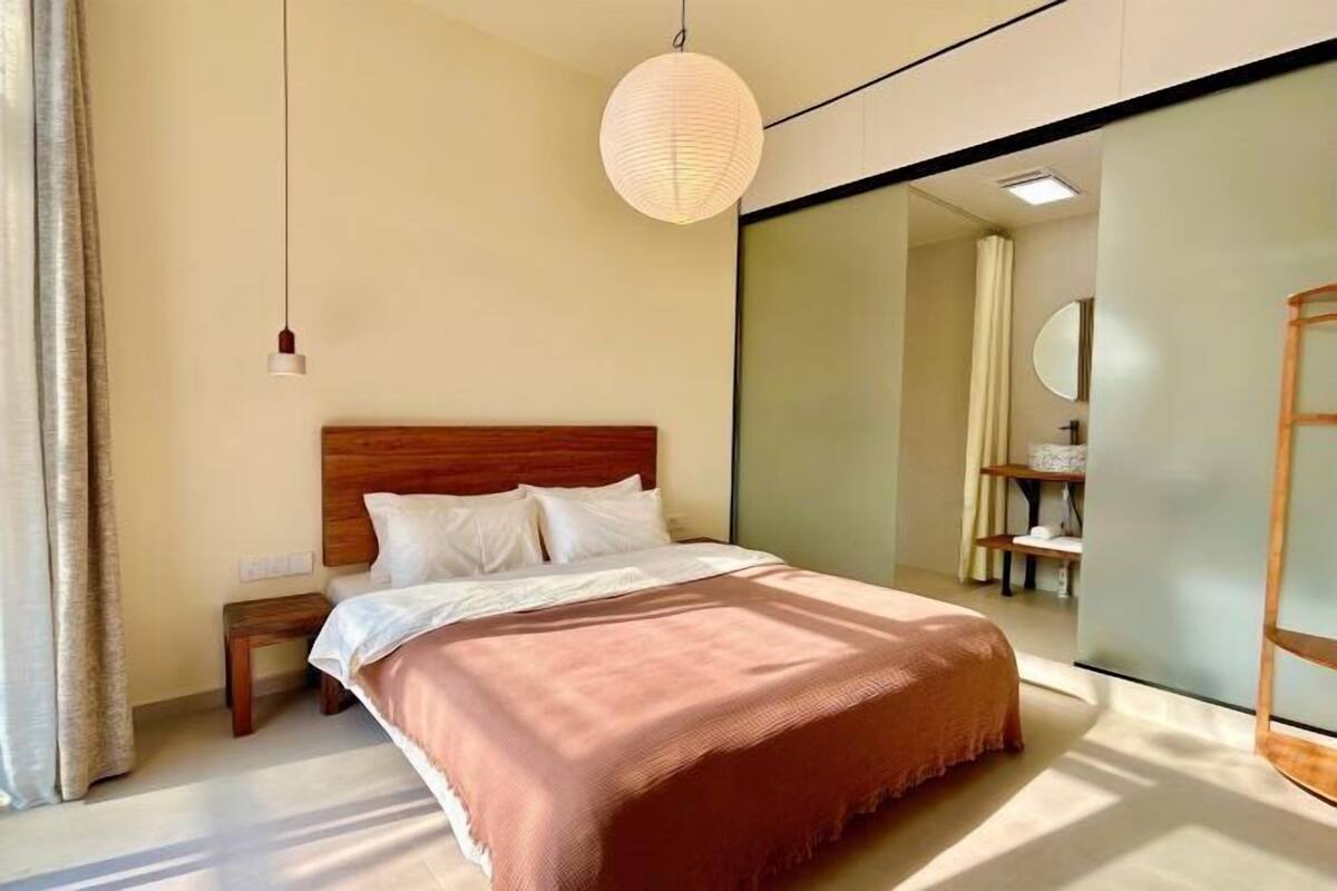 Een moderne slaapkamer met een houten hoofdbord, een groot bed met een roze deken, een nachtkastje en een hanglamp.