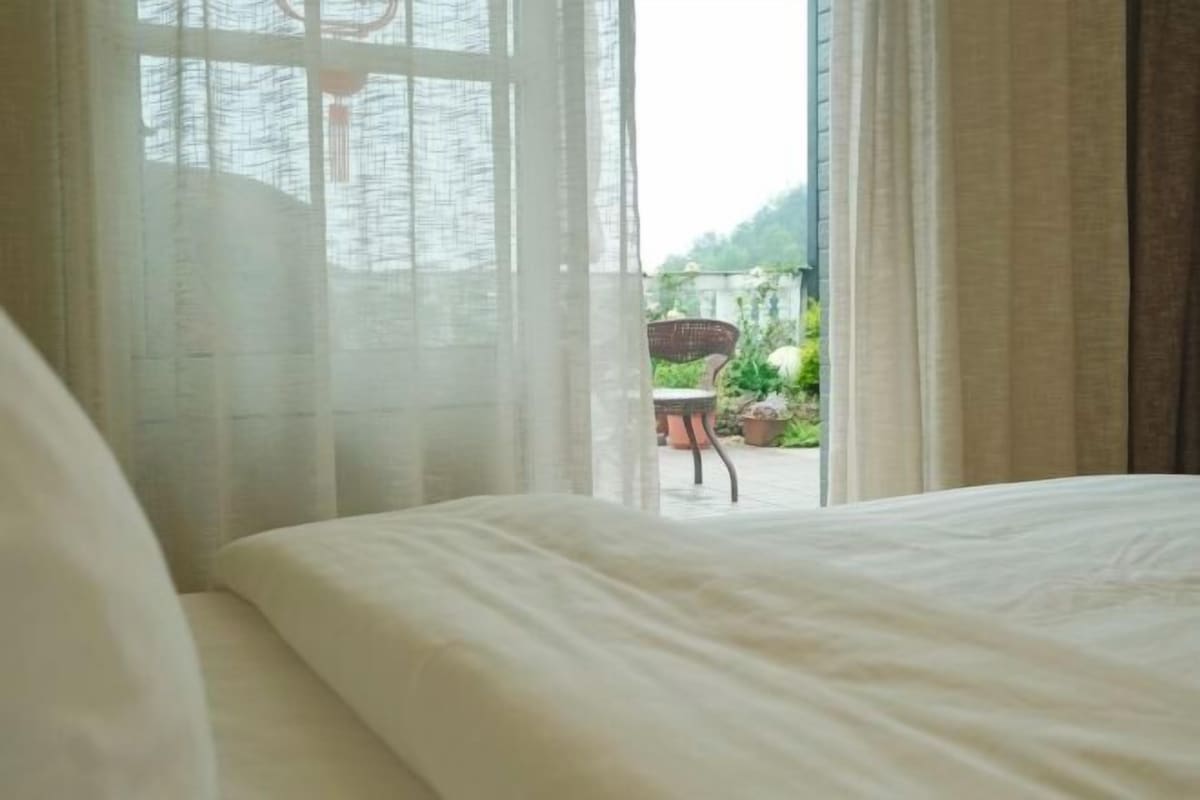 A bedroom with a bed, a window with sheer curtains, and a view of an outdoor area with a chair and potted plants.