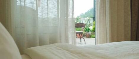 Un dormitorio con cama, ventana con cortinas translúcidas y vista a un espacio exterior con silla y plantas en macetas.