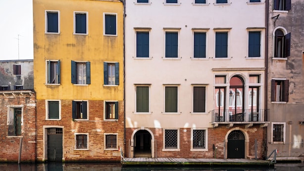 Un conjunto de edificios con varias ventanas y puertas, algunas con contraventanas, a lo largo de un canal que refleja las construcciones.