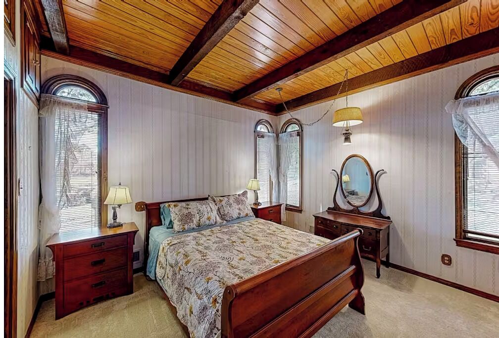 A bedroom with a wooden ceiling, two bedside tables, a bed with a floral bedspread, a mirror, and two windows with blinds.