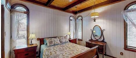 A bedroom with a wooden ceiling, two bedside tables, a bed with a floral bedspread, a mirror, and two windows with blinds.