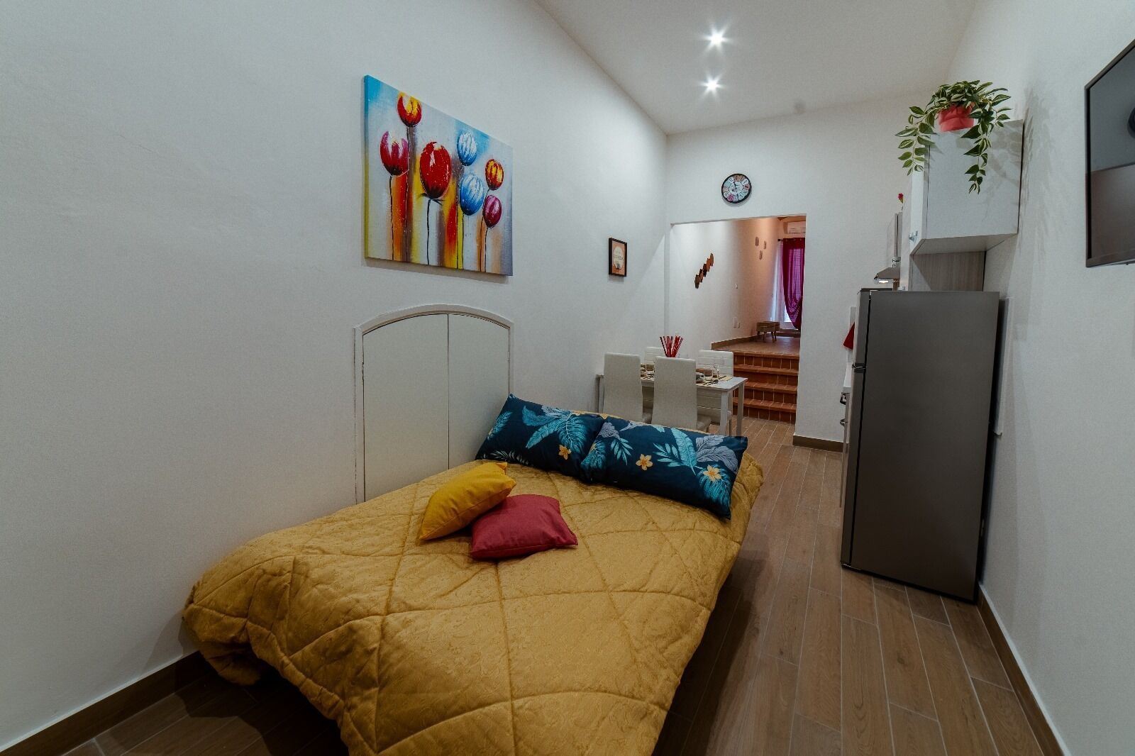 Un dormitorio con cama, escritorio, silla, refrigerador y un cuadro en la pared.