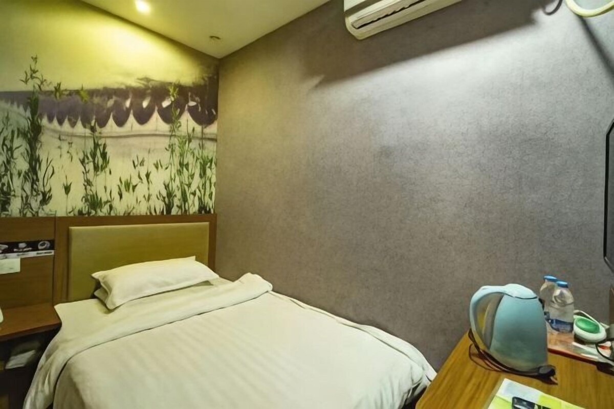 A hotel room with a bed, a desk with a chair, a water bottle, and a wall-mounted air conditioner.