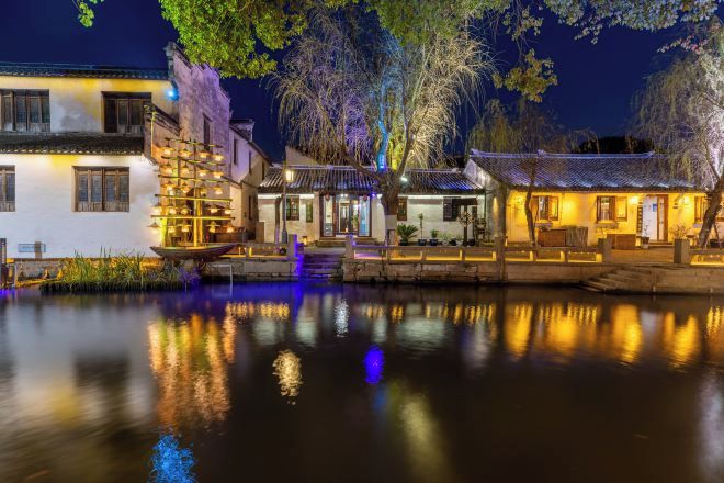 A traditional building with a water body in front, featuring a wooden structure with lights and a staircase leading to the entrance.