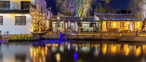 A traditional building with a water body in front, featuring a wooden structure with lights and a staircase leading to the entrance.