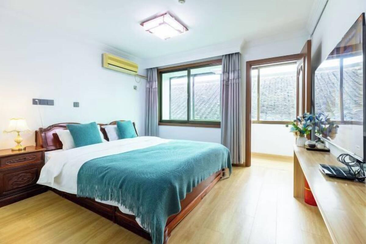 Una camera d'albergo con un letto in legno, un comodino con una lampada, un'ampia finestra con tende e una scrivania con una sedia.
