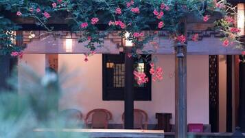 An outdoor seating area with a table and chairs, a pergola with flowering vines, and a traditional building facade in the background.