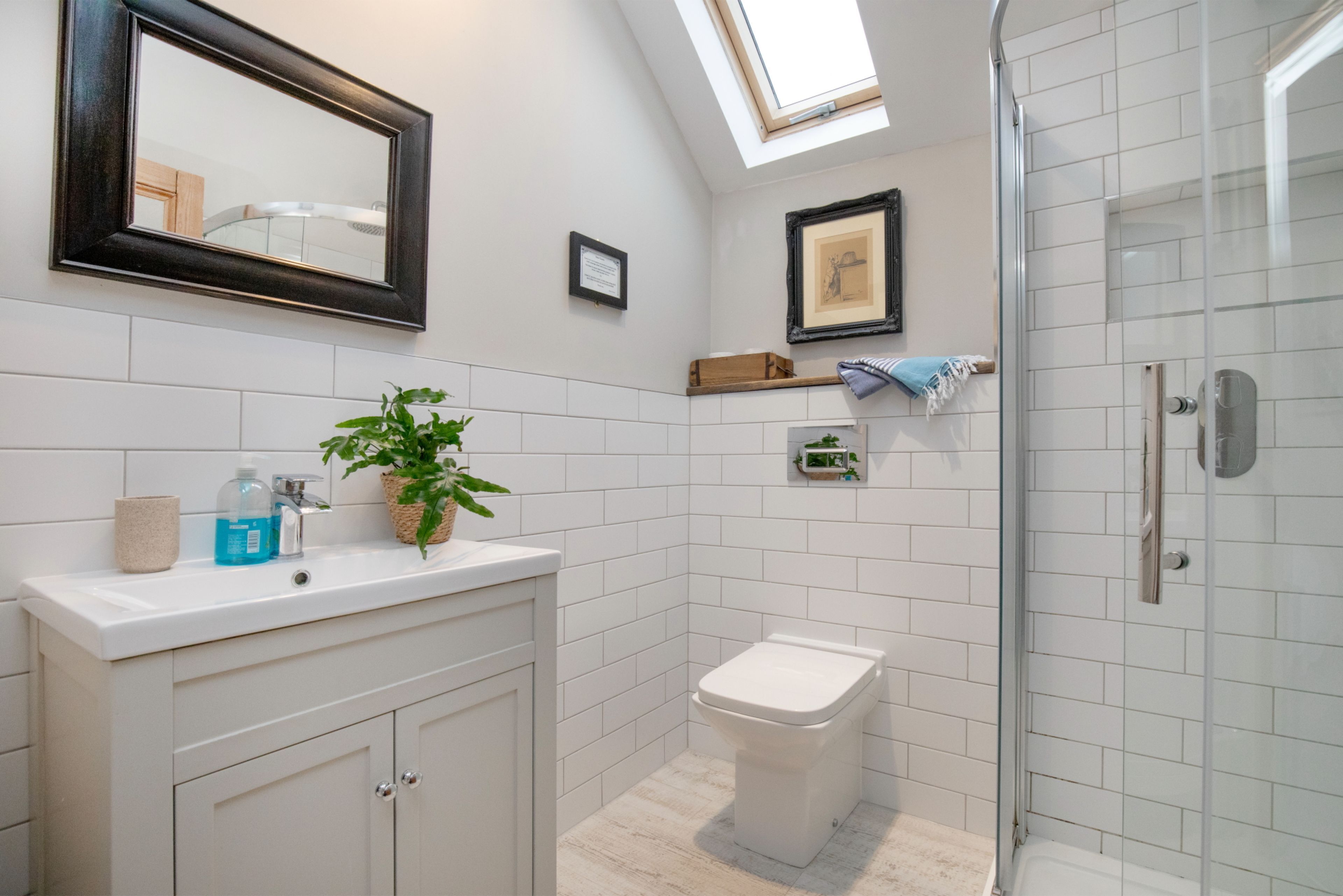 A bathroom with a skylight, white tiles, a toilet, a sink with toiletries, and a shower area.