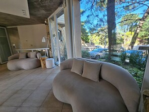 Ein modernes Wohnzimmer mit einer gebogenen Couch, einem kleinen Holzschemel und einer Glastür, die auf einen Balkon mit Blick auf Bäume und ein Auto führt.