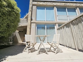 A two-story building with a balcony, a wooden deck with a table and chairs, and a large tree to the left.