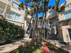 A multi-story building with outdoor staircases, a garden with flowers, and a clear blue sky.