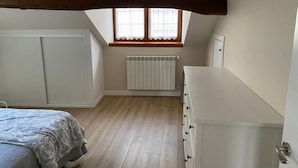 A bedroom with a sloped ceiling, a radiator, a window with white curtains, and a bed with a patterned blanket. - Loli's Attic (Luarca)
