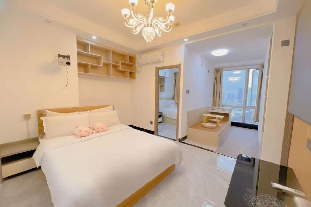 A modern hotel room with a large bed, a chandelier, a built-in TV, and a view of the city through large windows.