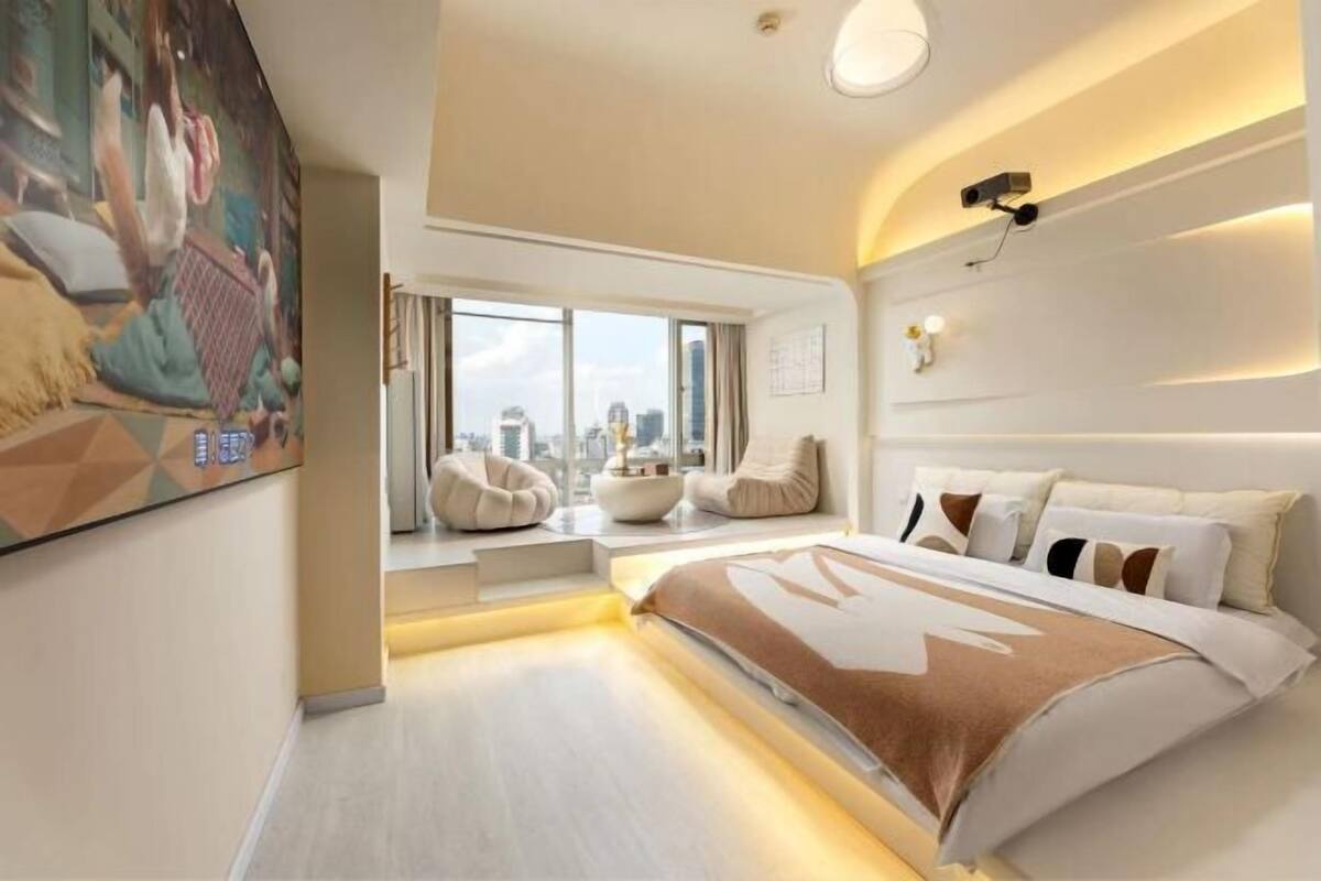 A modern hotel room with a large bed, a city view, a painting on the wall, and a projector on the ceiling.