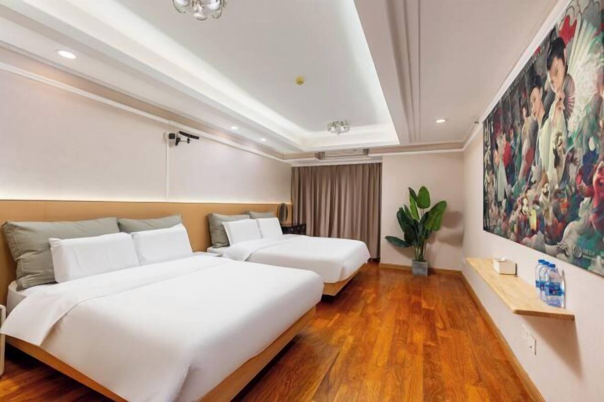 A hotel room with two beds, a large painting on the wall, a potted plant, and a wooden shelf with a water bottle.