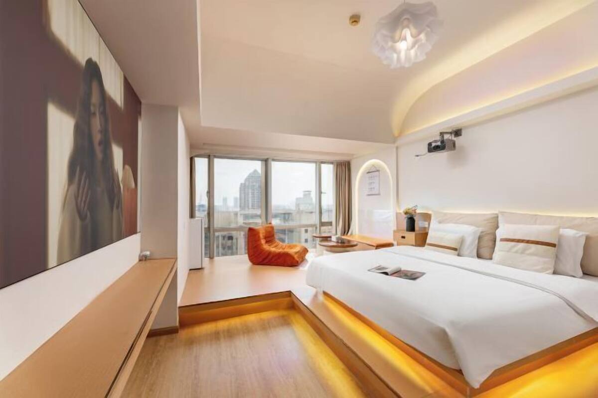 A modern hotel room with a large bed, a city view, a red chair, and a large painting on the wall.