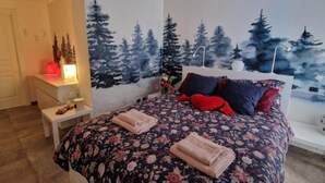 A bedroom with a floral bedspread, a white nightstand with a lamp, and a wall mural of trees. - The blue spruce room (Roma)
