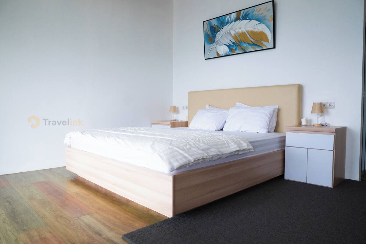 A modern hotel room with a wooden bed, white linens, a nightstand with a lamp, and a painting on the wall.