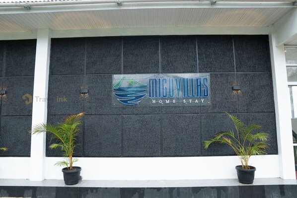 Fachada de edificio con un letrero que dice "MIGUVILAS HOME STAY" y dos macetas con plantas a cada lado.