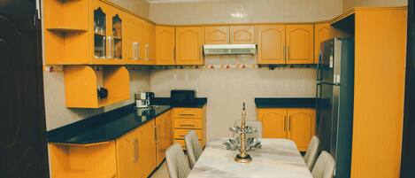 Una cocina con armarios de madera, encimera negra, una mesa de comedor con sillas y un refrigerador.